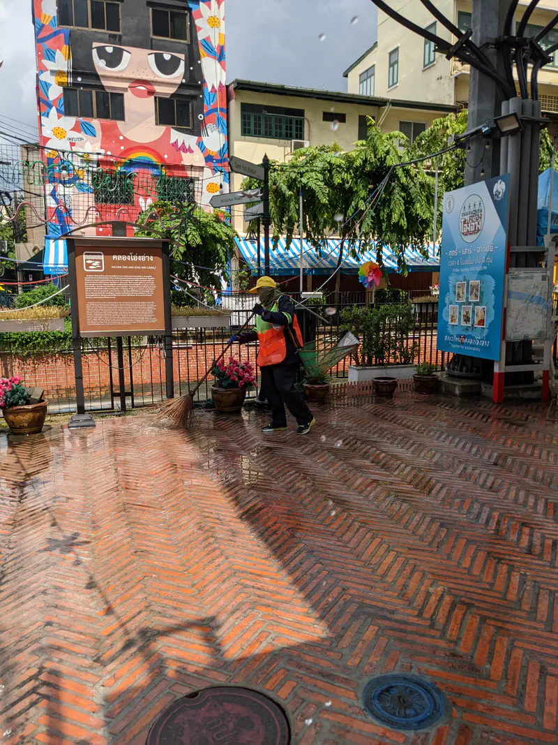 Street cleaner in uniform sweeping a wet brick pavement with a broom in front of a building with a large mural of a girl wearing a black hat and rainbow motifs.
