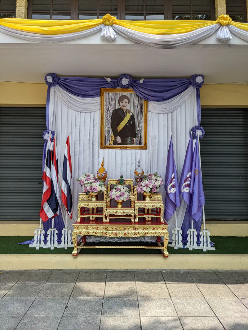 Large framed portrait of the King of Thailand on a golden pedestal outside a modern building.