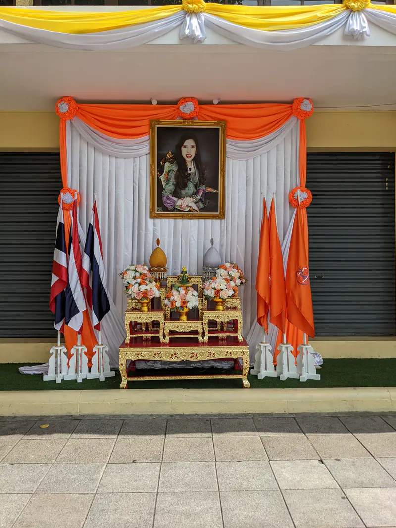 Street shrine with portrait of a Thai royal woman, orange drapes, flowers, and national flags in Bangkok.
