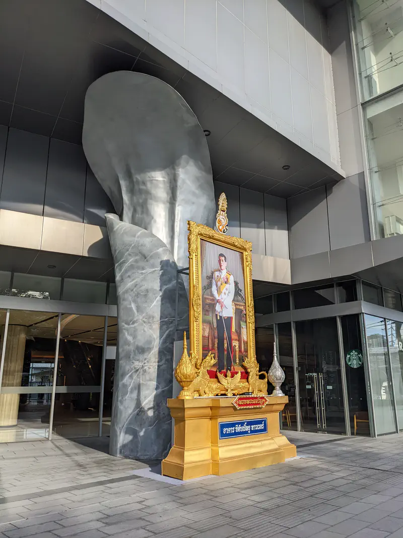 Large framed portrait of the King of Thailand on a golden pedestal outside a modern building.