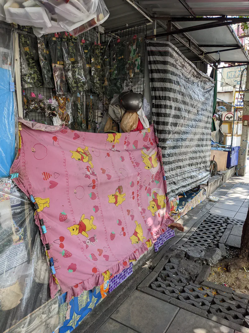 Street stall in Bangkok covered with tarps and a pink Winnie the Pooh blanket, selling camouflage clothing and accessories.
