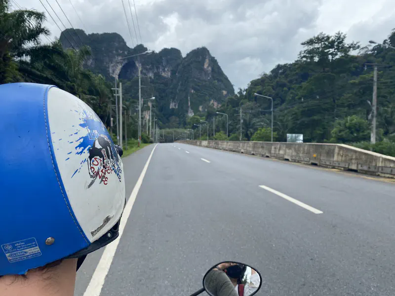 View from the back of a motorbike rider wearing a blue helmet, traveling along a highway flanked by limestone mountains and palm trees.