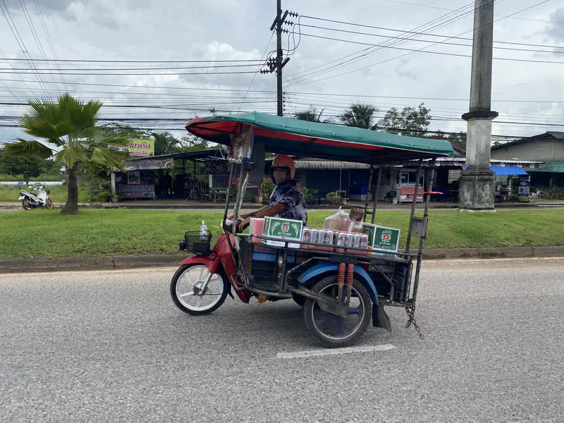 Man riding a motorbike with a sidecar loaded with boxes of drinks on a road with shops in the background.