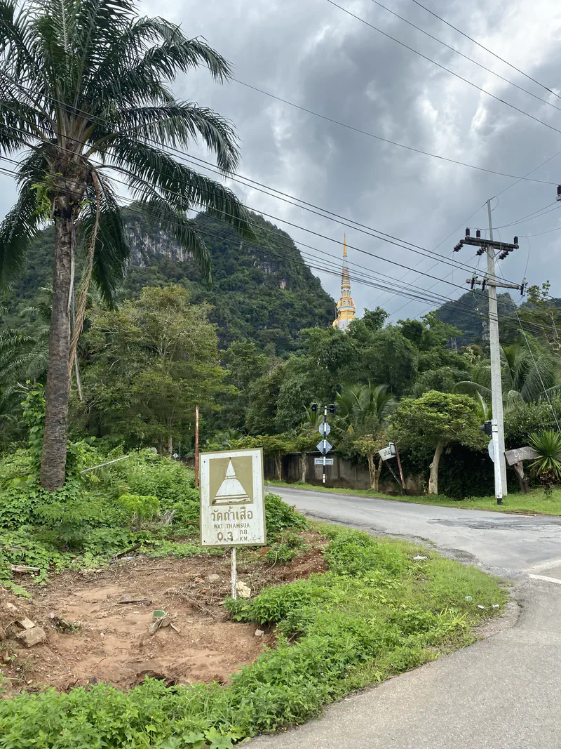 Roadside view with a sign pointing to Wat Thamsua, 0.3 km away, with a golden temple spire visible among green hills under a cloudy sky.