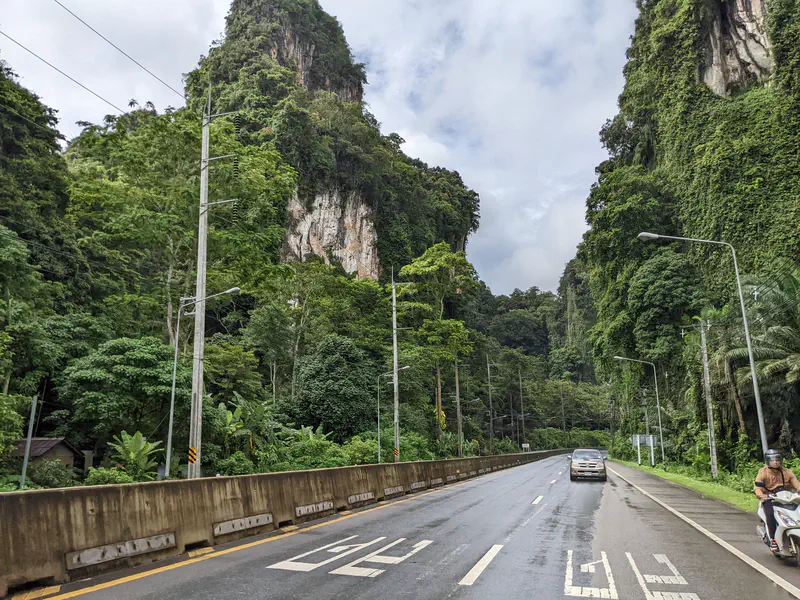 Highway cutting through towering limestone cliffs covered in dense green vegetation, with cars and motorbikes passing by.