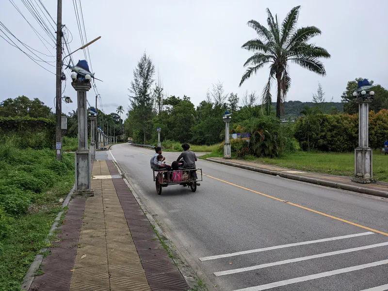 Two people riding in a motorbike sidecar along a quiet road lined with decorative lamp posts and palm trees.
