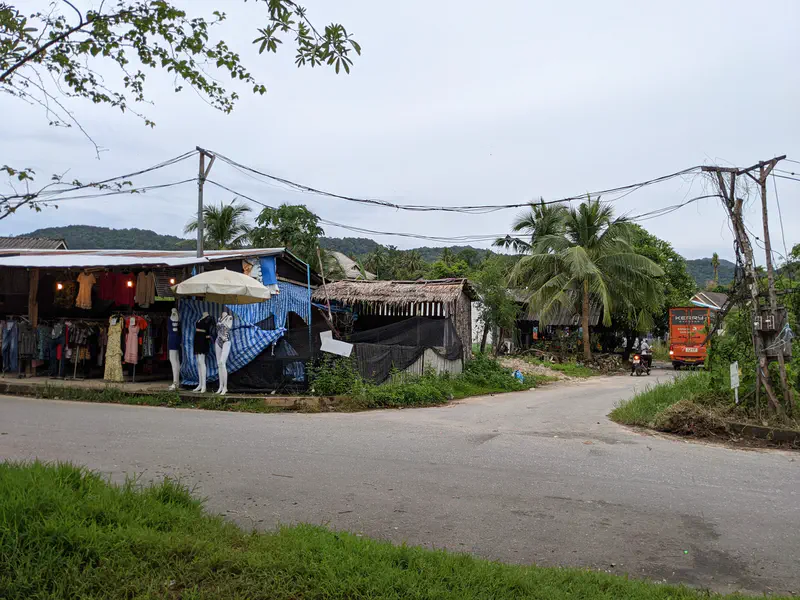 Small roadside market stall selling clothes with mannequins on display, next to a narrow road with a motorbike and bus passing by.