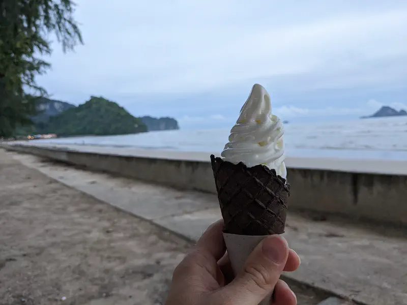 Hand holding a soft-serve ice cream cone with a beach and ocean view in the background.