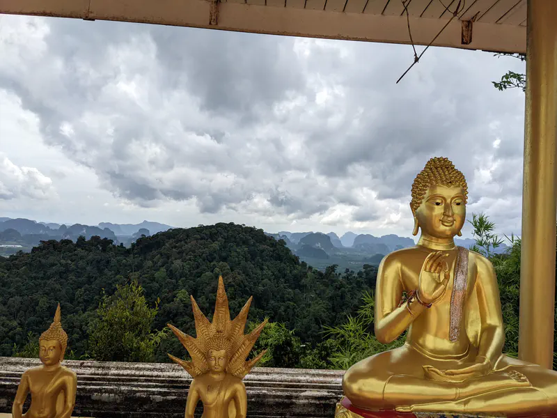 Golden Buddha statues at a mountain viewpoint overlooking green hills and dramatic limestone cliffs with cloudy skies.