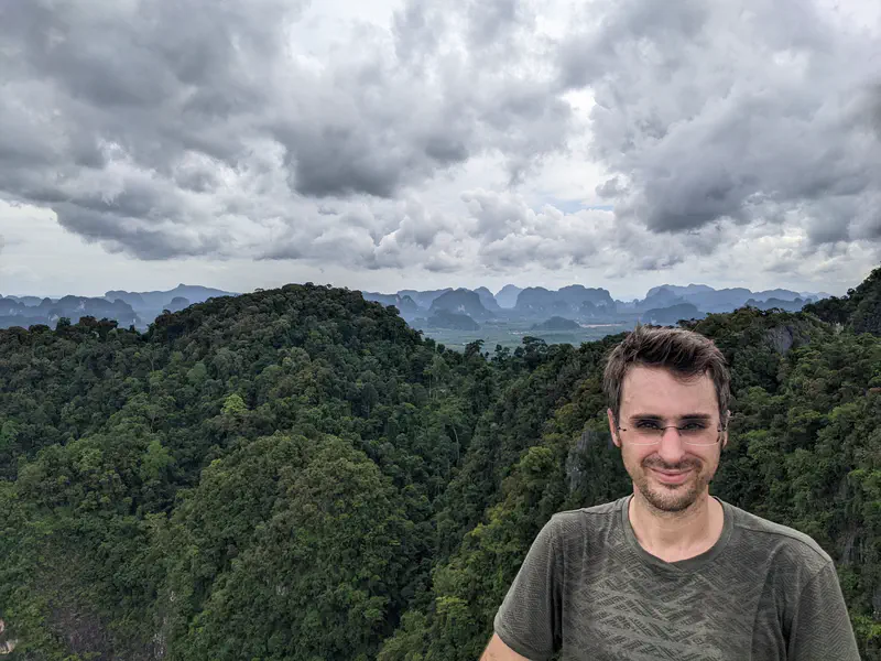 Man standing on a viewpoint with lush green forested hills and distant limestone mountains under a cloudy sky.