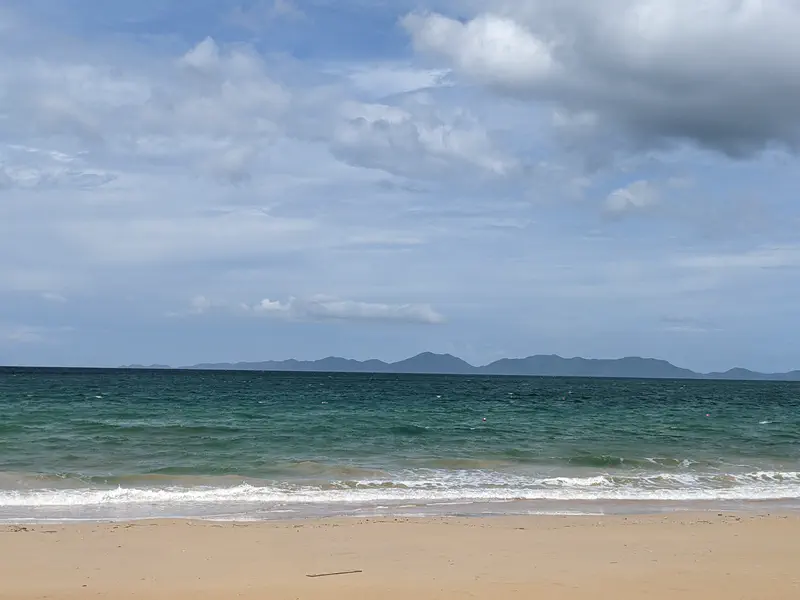 Sandy beach with gentle waves and a view of distant islands across the turquoise sea under a partly cloudy sky.