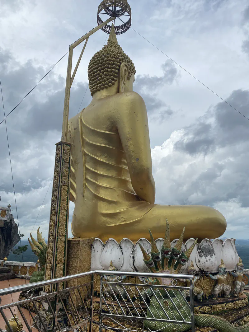 Large golden Buddha statue viewed from behind, seated on a decorated lotus pedestal with naga sculptures, under a cloudy sky.