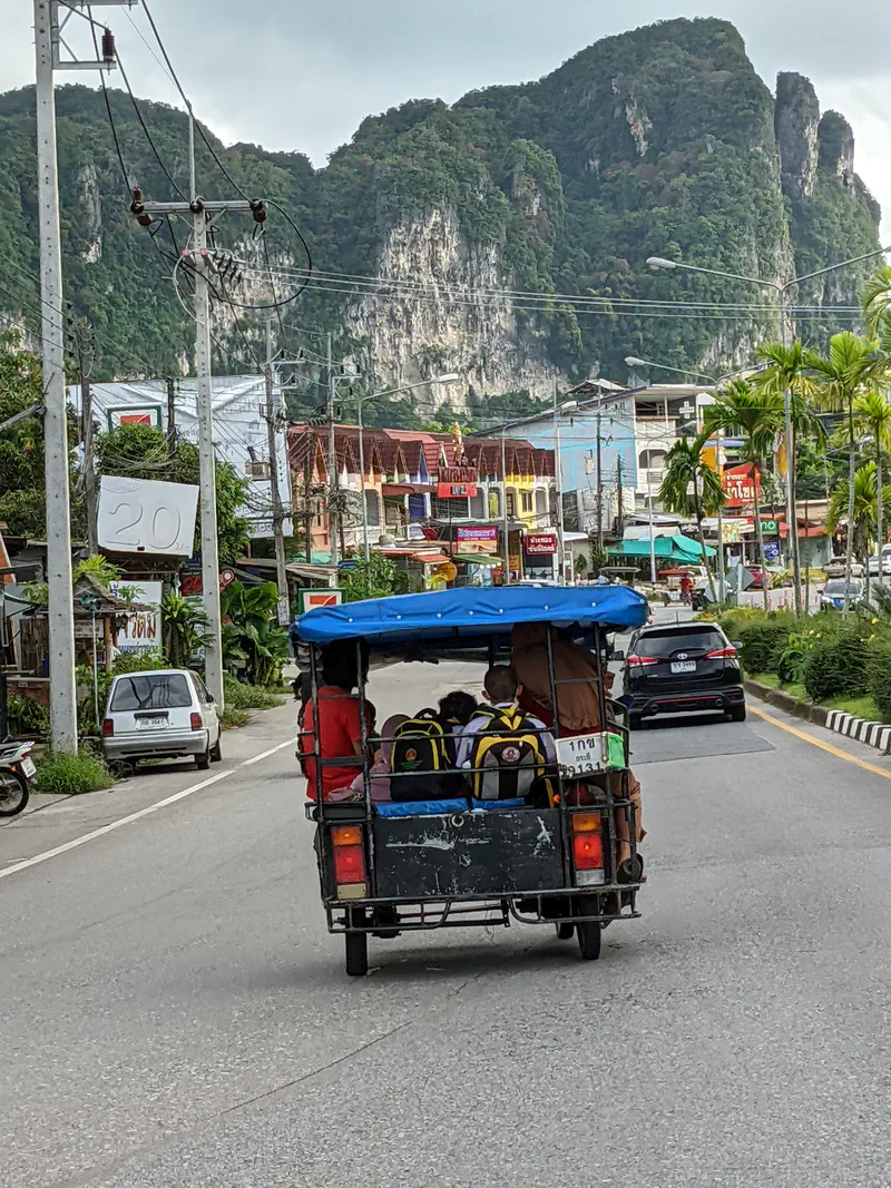Motorbike sidecar taxi carrying schoolchildren with backpacks through a town with limestone cliffs in the background.