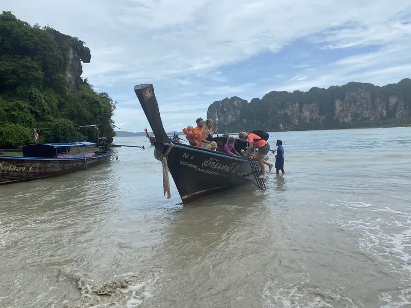 Tourists boarding a wooden long-tail boat at the shore with limestone cliffs in the background.