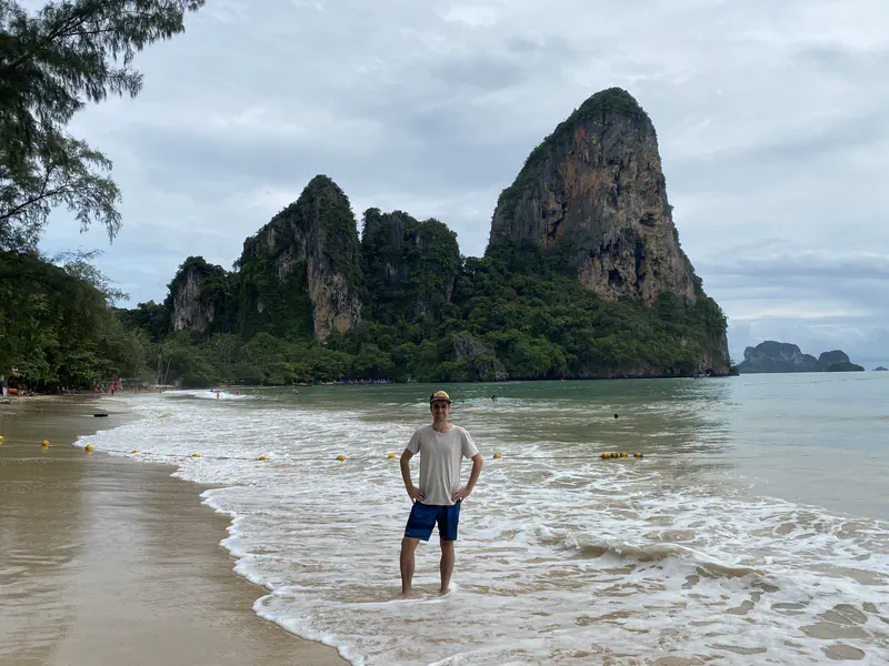 Person standing on a sandy beach with waves and tall limestone cliffs covered in greenery behind.