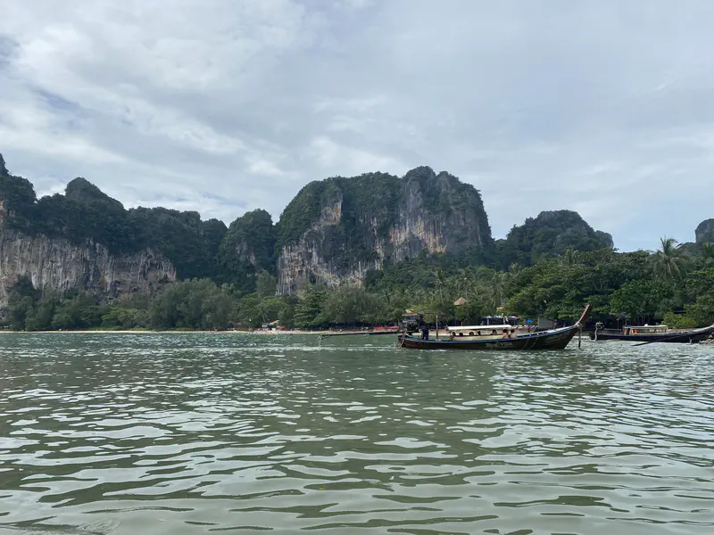 Long-tail boats floating on calm water with a backdrop of limestone cliffs and lush green trees under a partly cloudy sky.