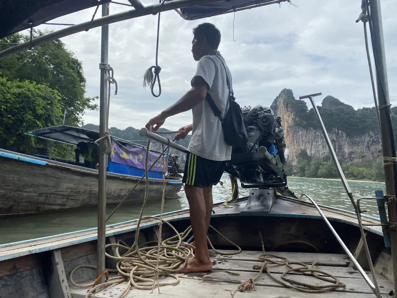 Boatman standing barefoot at the back of a long-tail boat steering near limestone cliffs and other boats on the water.