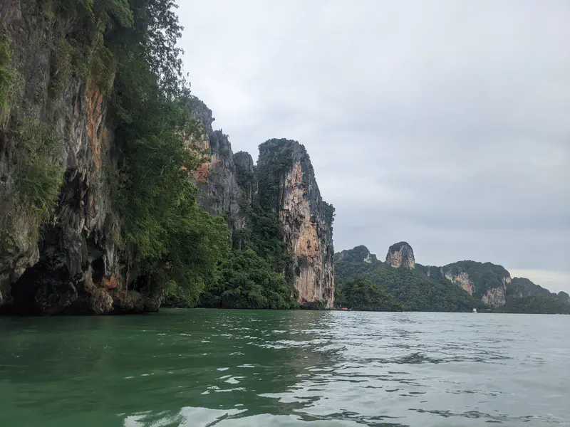 Limestone cliffs with dense greenery rising from calm green water under an overcast sky.