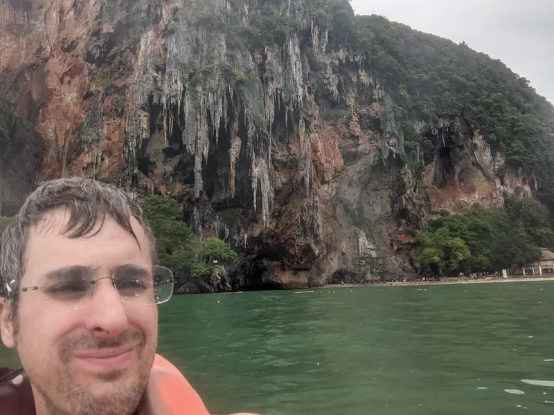 Person wearing glasses and a life jacket in the water with towering limestone cliffs and a cave in the background.