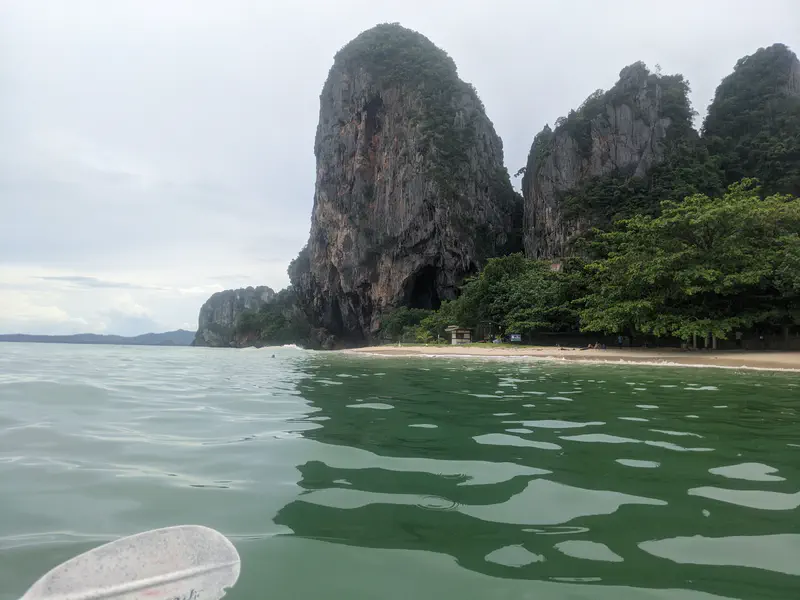 View from the water of a sandy beach backed by towering limestone cliffs and lush green trees under a cloudy sky.