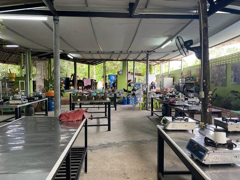 Open-air cooking school with multiple cooking stations, gas stoves, and tables set up under a roof with green walls and kitchen equipment.