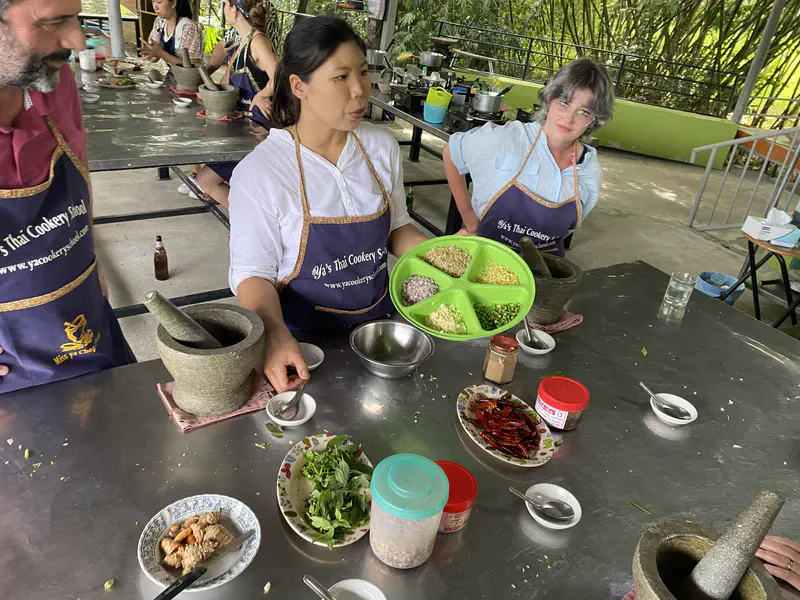 Instructor at a Thai cookery school holding a tray of chopped ingredients while students in aprons watch at a cooking station with spices and herbs on the table.