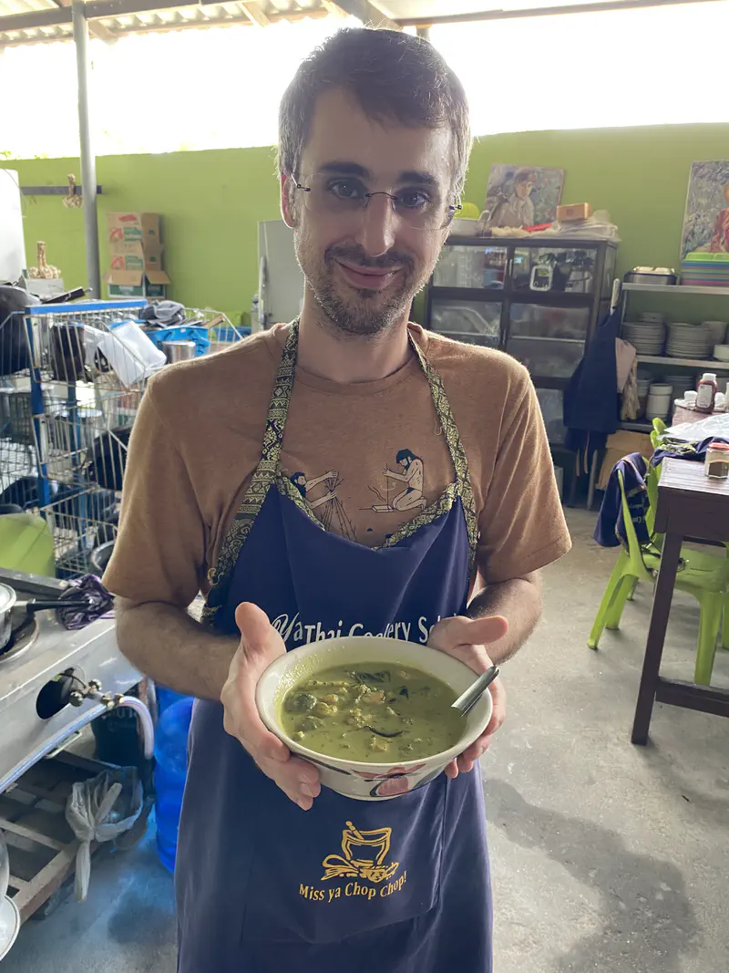 Person in a cooking school apron smiling and holding a bowl of freshly made Thai green curry.