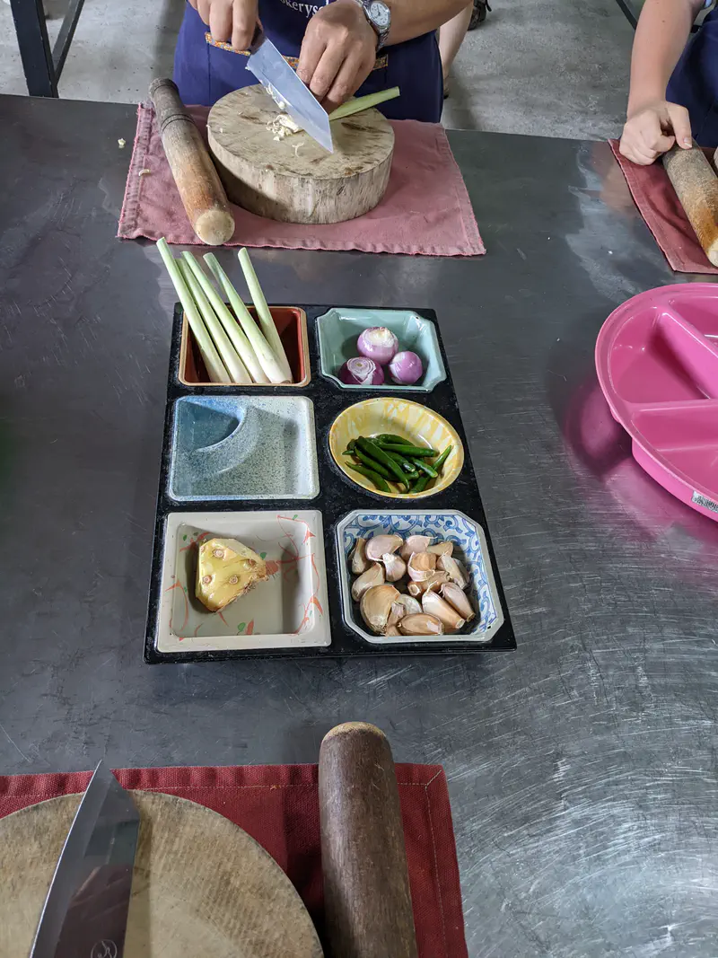Cooking class setup with trays of fresh ingredients including lemongrass, shallots, green chilies, galangal, and garlic, while someone chops on a wooden board.