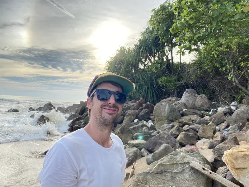 Person in sunglasses and a cap smiling on a rocky beach with waves crashing and the sun setting behind trees.