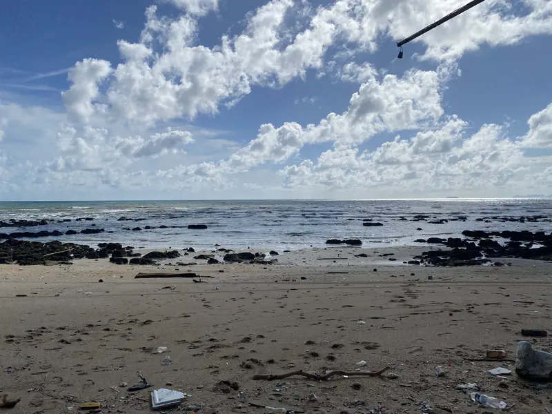 Beach view with scattered rocks, waves rolling in under a partly cloudy sky, and litter visible on the sand.
