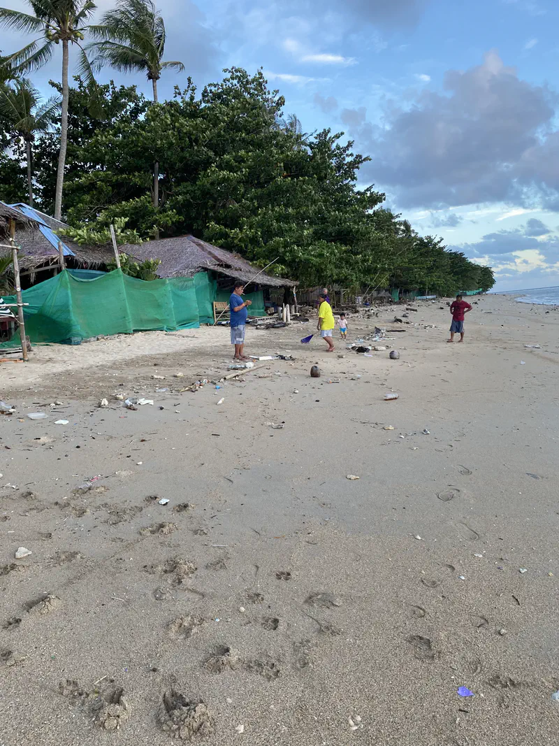 People standing on a sandy beach near simple huts and palm trees, with noticeable litter scattered along the shore.