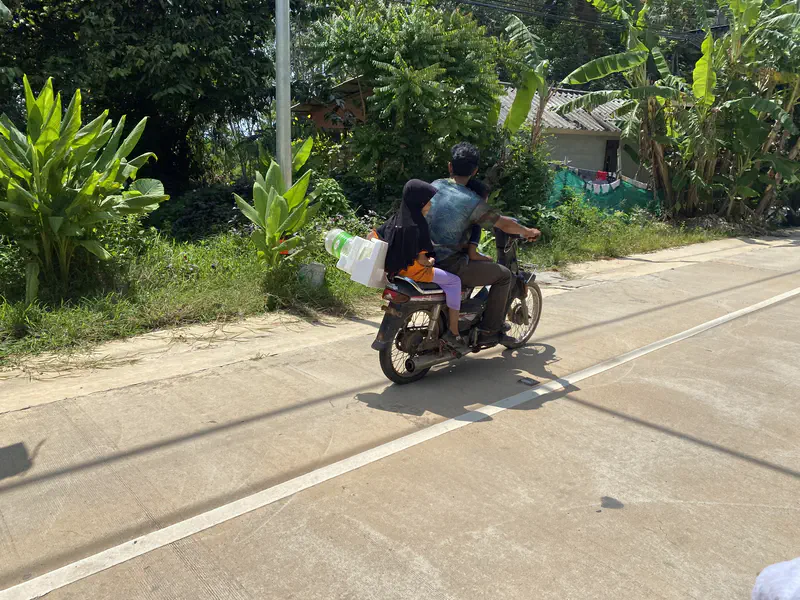 Man riding a motorcycle with a child seated on the back, carrying plastic containers, on a sunny rural road with greenery.