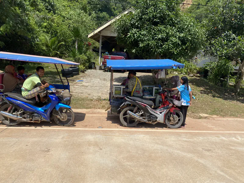 Street food vendors operating from motorbikes with blue canopies, one man smiling on a blue motorbike while others sell food to a woman by the roadside.