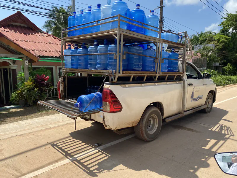 White pickup truck loaded with large blue water containers stacked on metal racks, parked on a sunny street near houses and greenery.