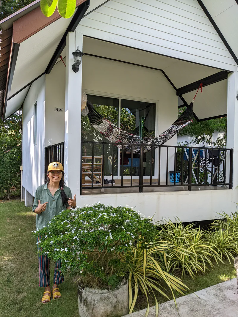 Person standing and smiling in front of a small white bungalow with a hammock on the porch surrounded by greenery.