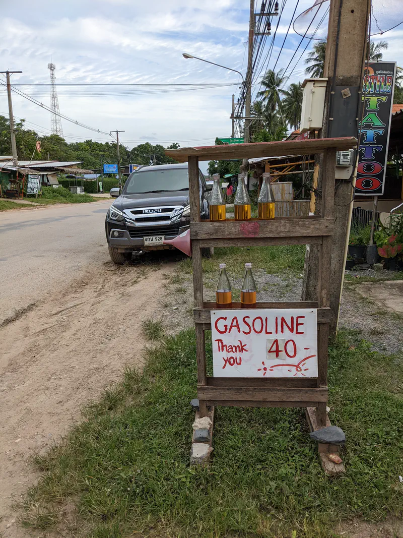 Roadside wooden stand with glass bottles of gasoline for sale, marked with a handwritten sign reading 'GASOLINE 40 Thank you'.