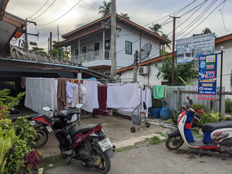 Laundry service shop with clothes and bedding hanging to dry outside on racks, with motorbikes parked in front and signs in Thai and English.