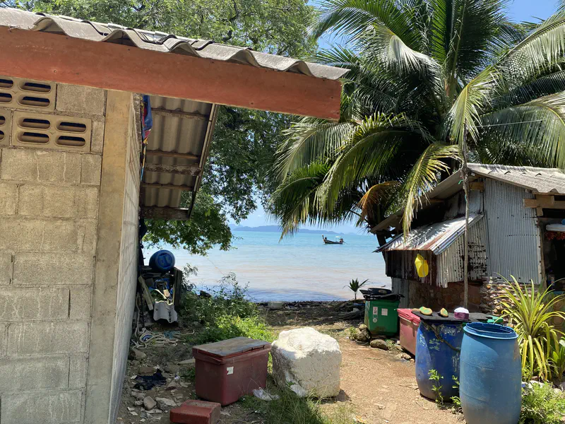 Seaside village view with simple houses, palm trees, and blue barrels, looking out to the sea with a longtail boat in the distance.