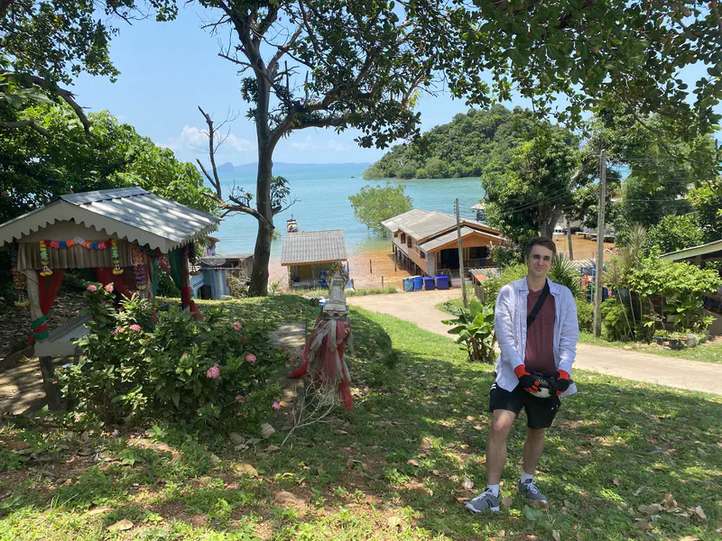 Person standing on a grassy hill overlooking a coastal village with houses, trees, and the turquoise sea in the background.