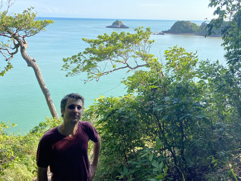 Person standing on a lush hillside with a view of turquoise sea and small islands in the background, partially framed by trees.