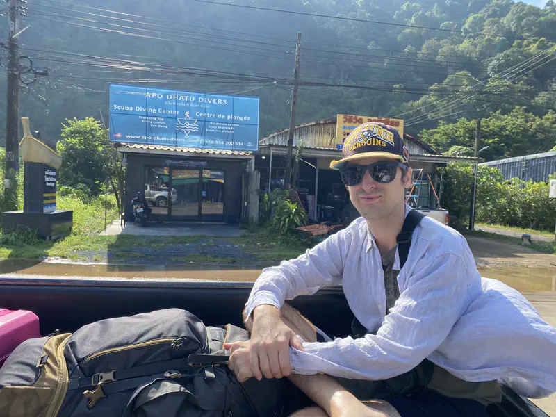 Person wearing sunglasses and a cap sitting in the back of a vehicle with a backpack, in front of a scuba diving center sign and lush green hills.