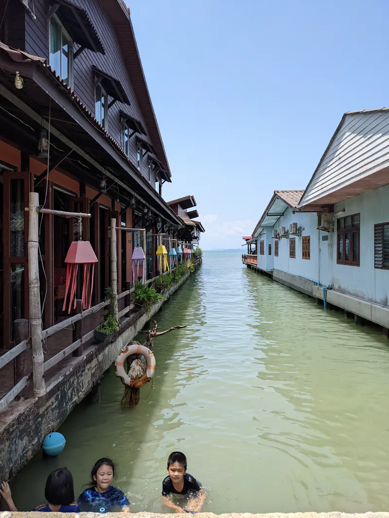 Children swimming in a narrow canal between stilt houses by the sea on a sunny day.