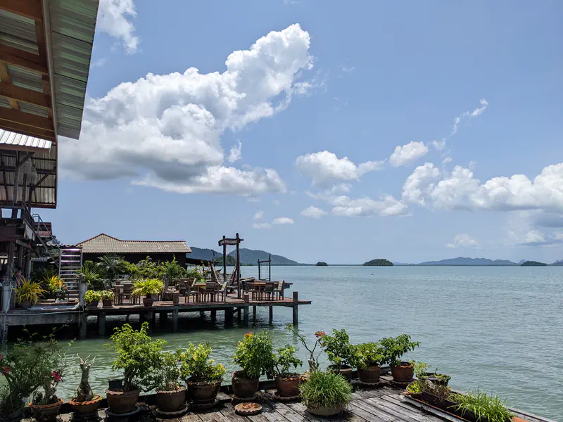 Wooden deck with potted plants and outdoor seating overlooking calm turquoise sea with islands in the distance under a partly cloudy sky.