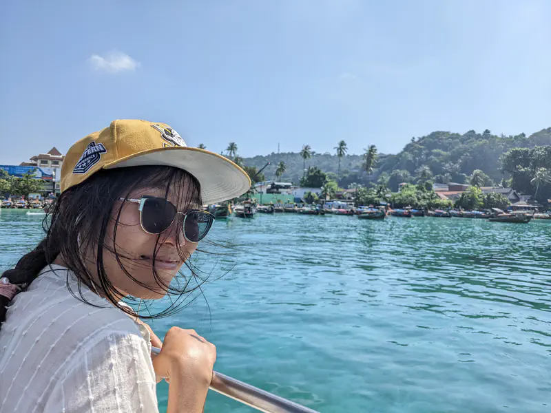 Person wearing a cap and sunglasses smiling on a boat with a view of turquoise water and a coastal village in the background.