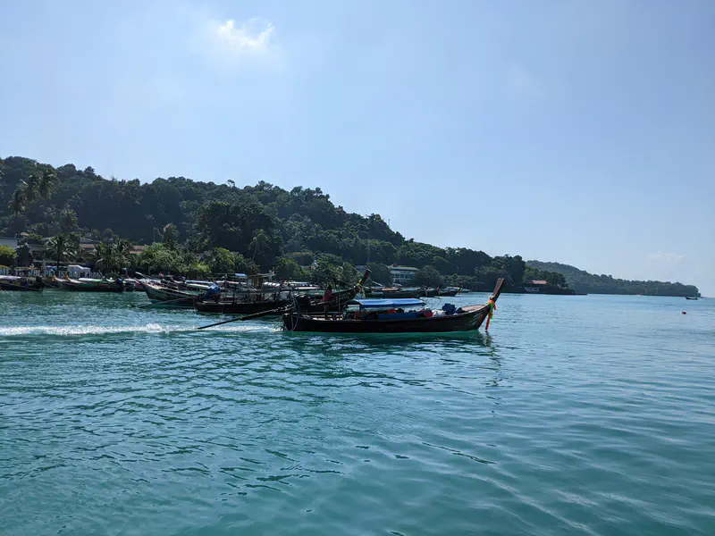 Traditional long-tail boats floating on turquoise water near a forested coastline under a clear sky.