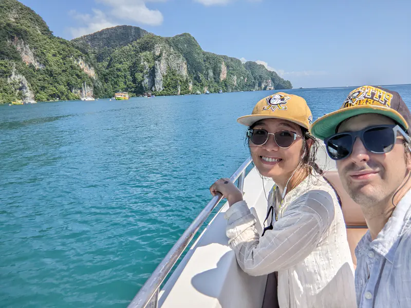 Two people smiling on a boat with turquoise water and green cliffs in the background on a sunny day.