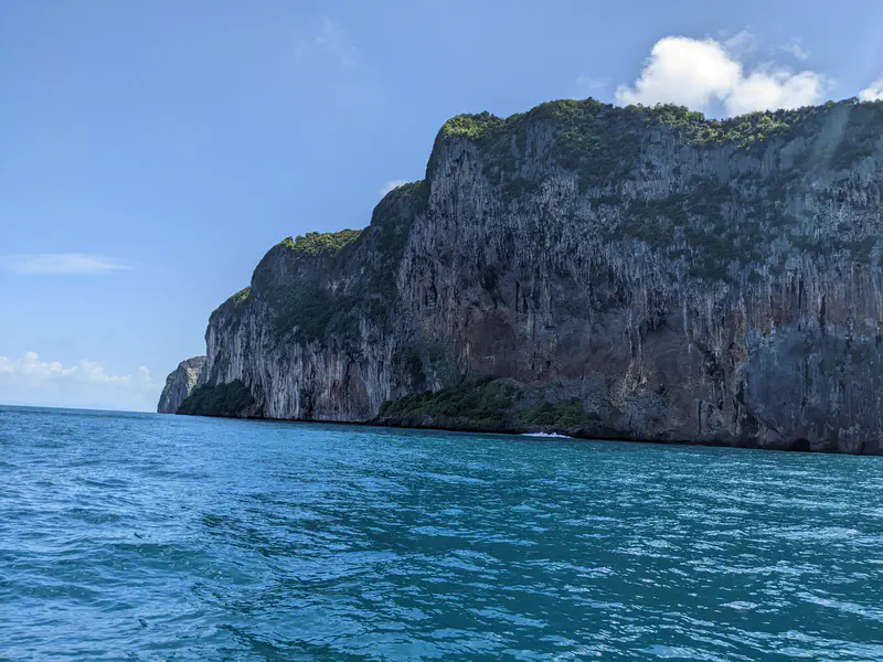 Massive rocky cliff covered with greenery rising from turquoise sea under a blue sky with light clouds.