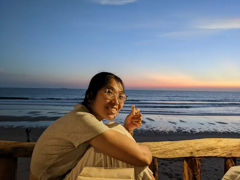 Person smiling while sitting by a wooden railing at the beach during sunset, with the ocean and colorful sky in the background.