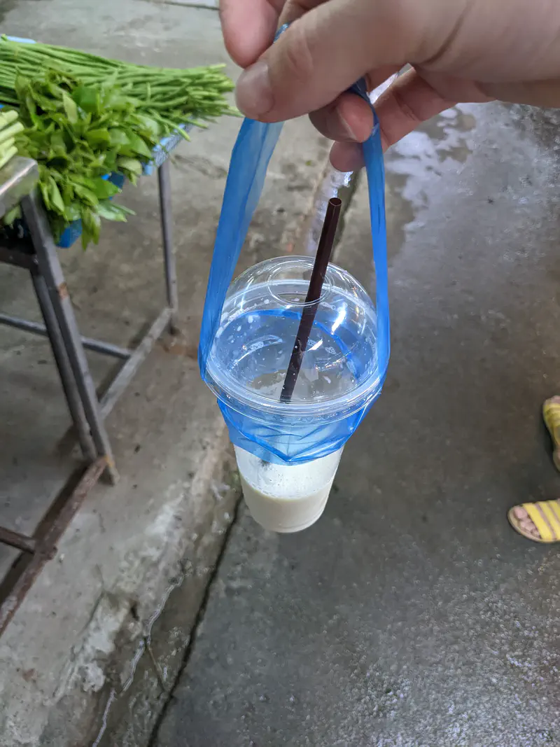 Hand holding a plastic cup of iced drink with a straw, carried in a blue plastic handle at a street market.