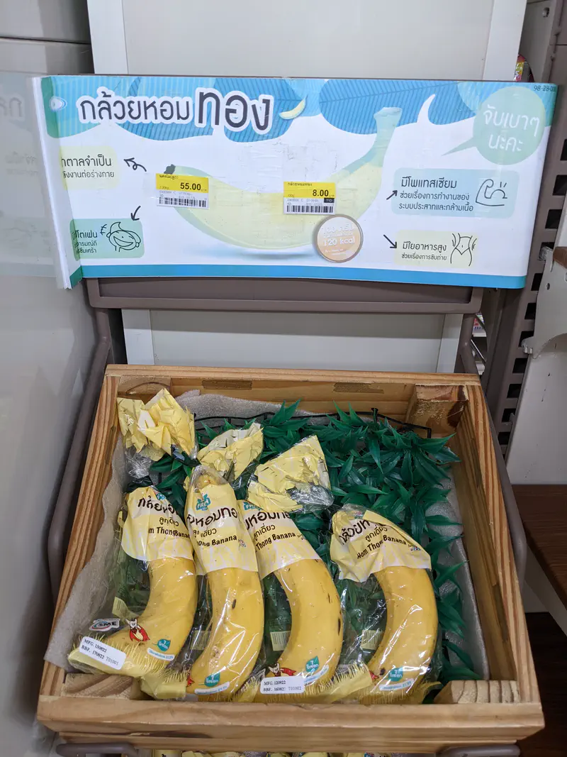 Packaged bananas labeled 'Hom Thong Banana' displayed in a wooden crate with a sign above showing nutritional information and pricing in Thai.